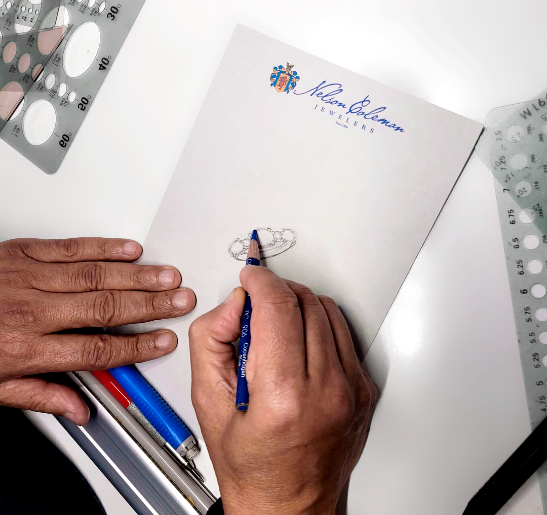 A desk type workspace with the left hand holding a paper while the right hand sketches a custom ring design. The paper has the Nelson Coleman Jewelers logo at the top.
