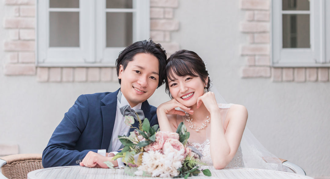 A bride and groom in wedding attire sitting at a table with flowers and smiling