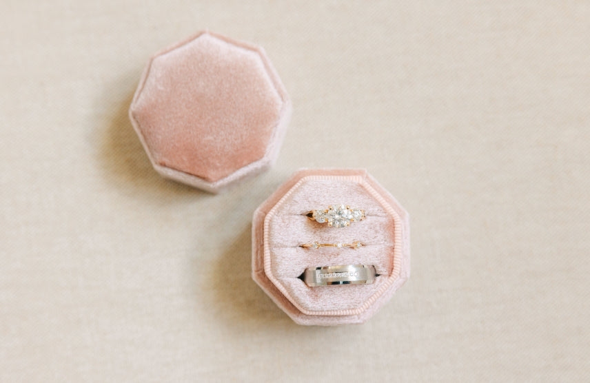 An octagonal pink velvet ring box with the lid off sitting next to it. Inside is a diamond engagement ring, a diamond wedding band, and a thick band channel set with diamonds.