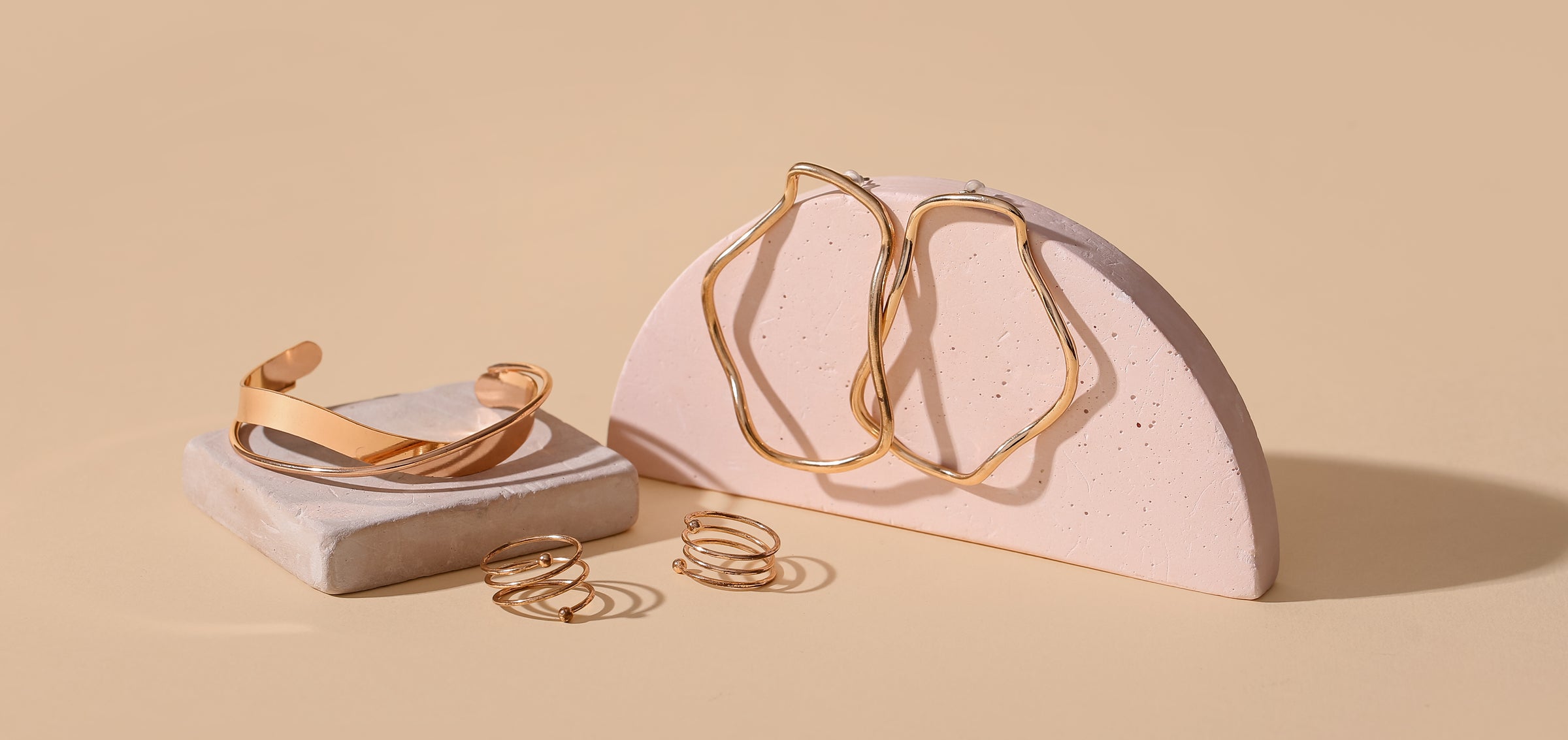 Gold hoop earrings on a pink stand, a gold bracelet on an off white square tile, and gold spiral rings with a beige background