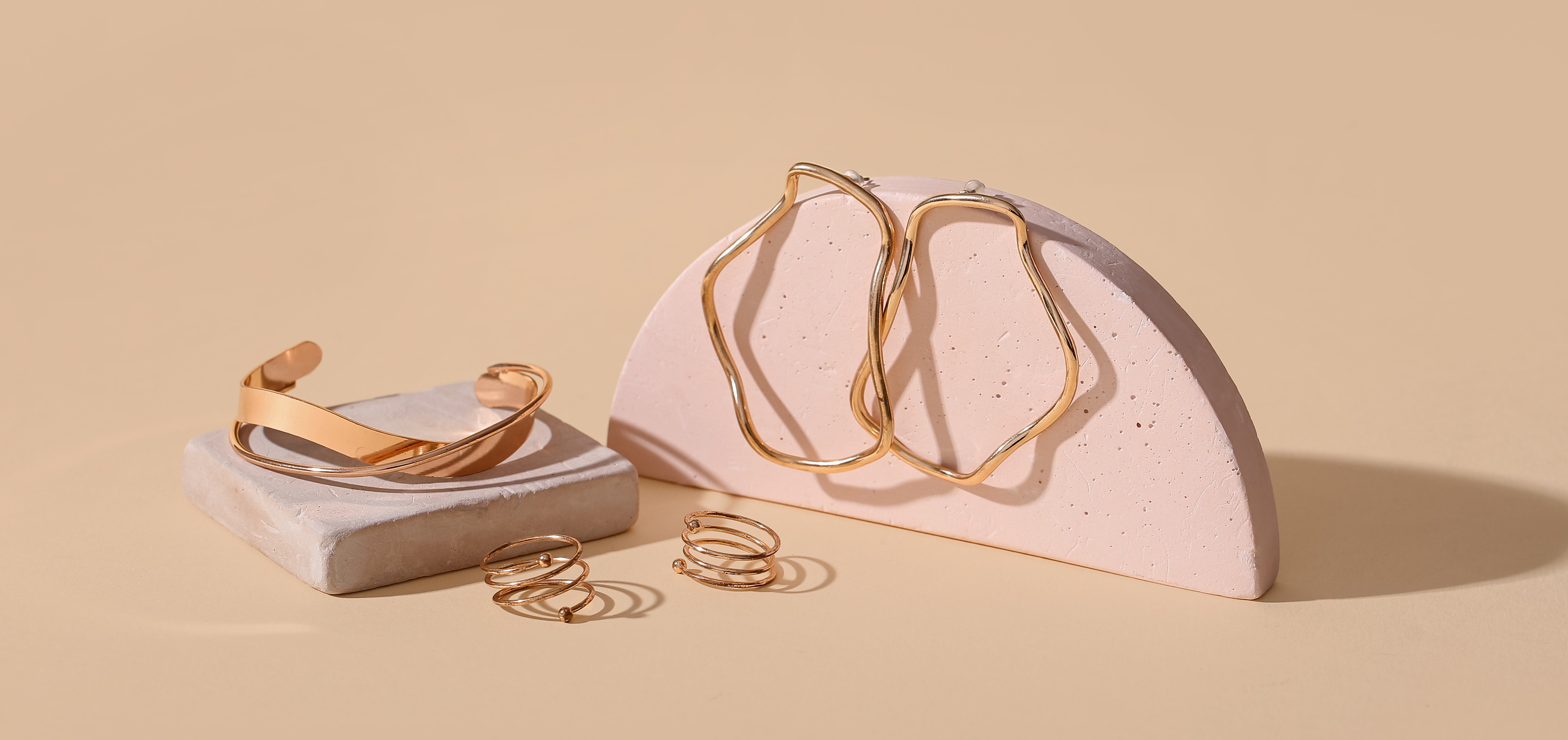 Gold hoop earrings on a pink stand, a gold bracelet on an off white square tile, and gold spiral rings with a beige background
