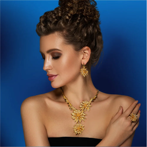 A stylish young woman wearing a gold flower jewelry set of earrings, a necklace, and a ring on a blue background