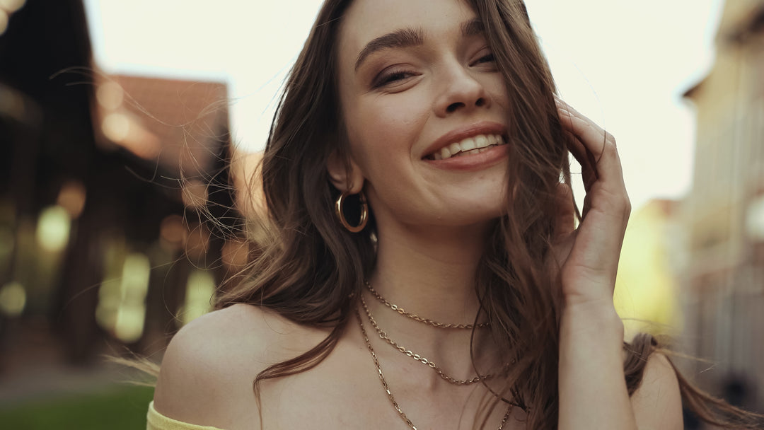 Woman with gold jewelry, long hair, and a yellow top smiling outdoors with a blurred background
