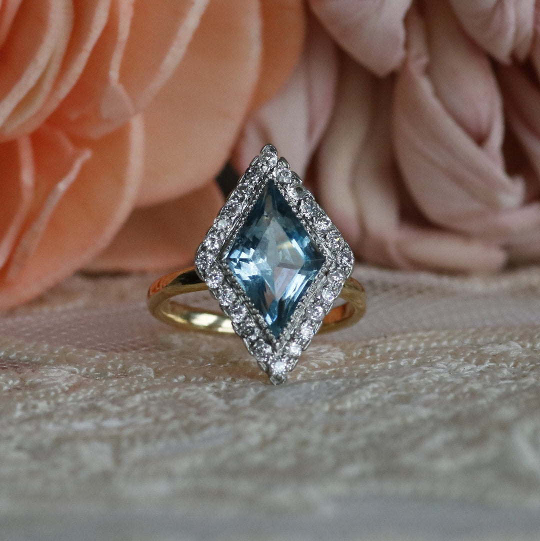 A diamond-cut blue topaz ring with a halo of small diamonds and a yellow gold band, displayed on a lace surface between peach and pink fabric flowers.
