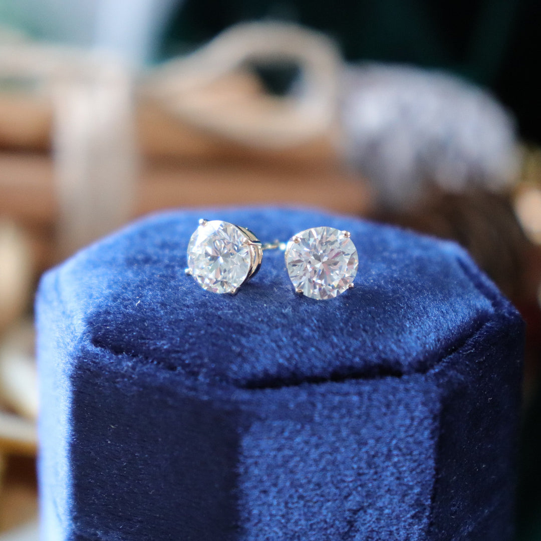 A pair of round-cut diamond stud earrings in a yellow gold setting, displayed on a dark blue velvet jewelry box with a warm, festive blurred background.