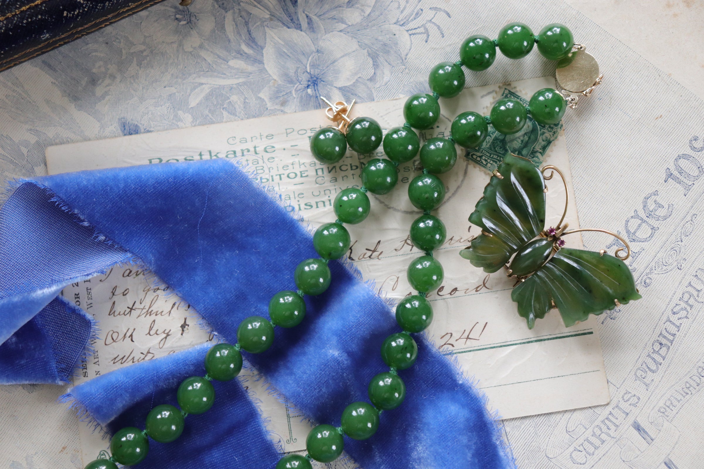 An estate green jade jewelry set including jade ball stud earrings, a jade bead necklace, and a carved jade butterfly brooch resting on a periwinkle velvet ribbon and vintage-style postcards.