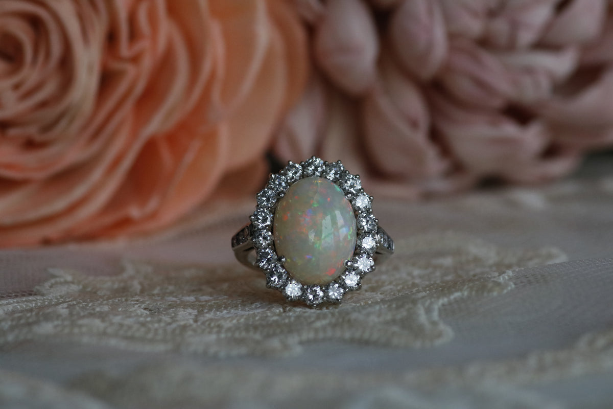 A close-up image of an estate white gold oval opal and diamond halo ring on white lace with pink flowers blurred in the background.