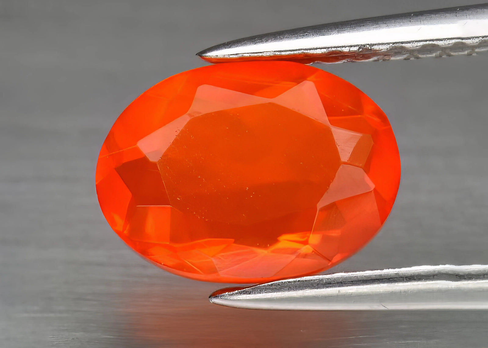 An oval-shaped faceted fire opal held by tweezers on a grey background.
