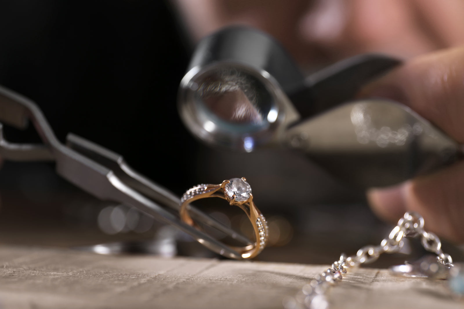A close-up image of a professional jewelry of a yellow gold diamond engagement ring being held with tweezers and examined using a jeweler's loupe.
