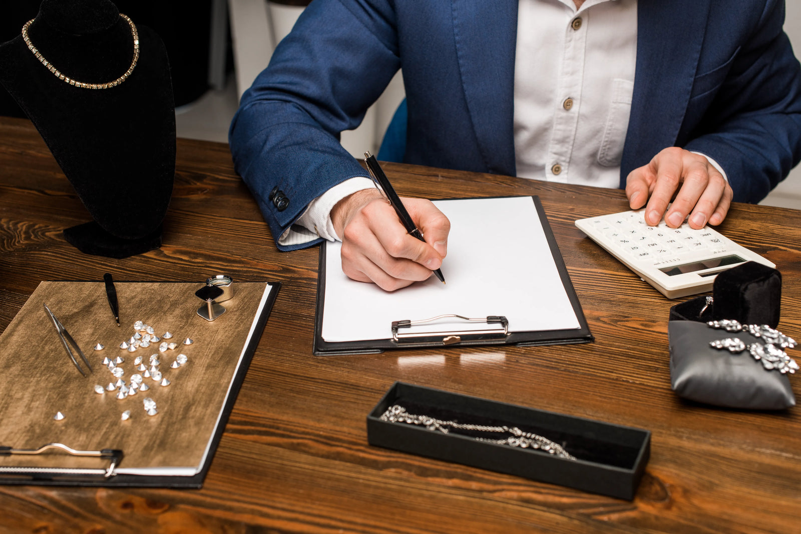 A close-up scene of a professional jewelry appraisal with a man making notes on a clipboard while using a calculator. On the desk near him is a clipboard with loose diamonds, tweezers, and a jeweler's loupe, as well as pieces of diamond jewelry.