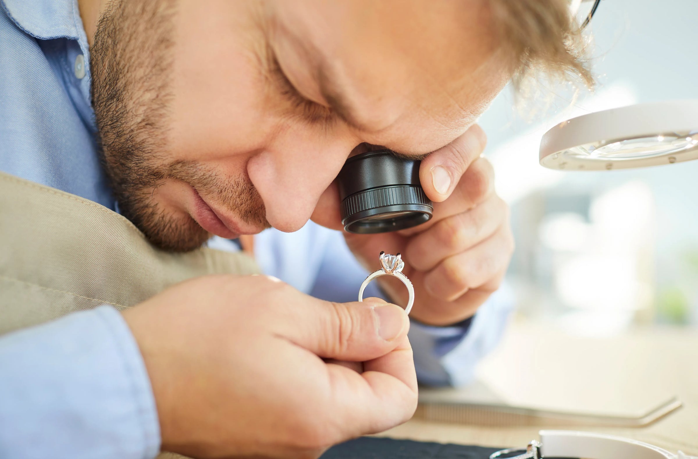 A close-up lifestyle shot of a male jeweler conducting a professional jewelry appraisal with a diamond engagement ring under a loupe microscope. The jeweler is wearing a light blue shirt and beige apron, with a magnifying lamp visible on the right against a bright, blurred background.