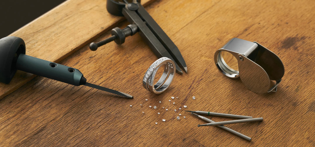 A wooden tabletop with a diamond cross over band, small loose diamonds, a jeweler's loupe and jewelry repair tools.