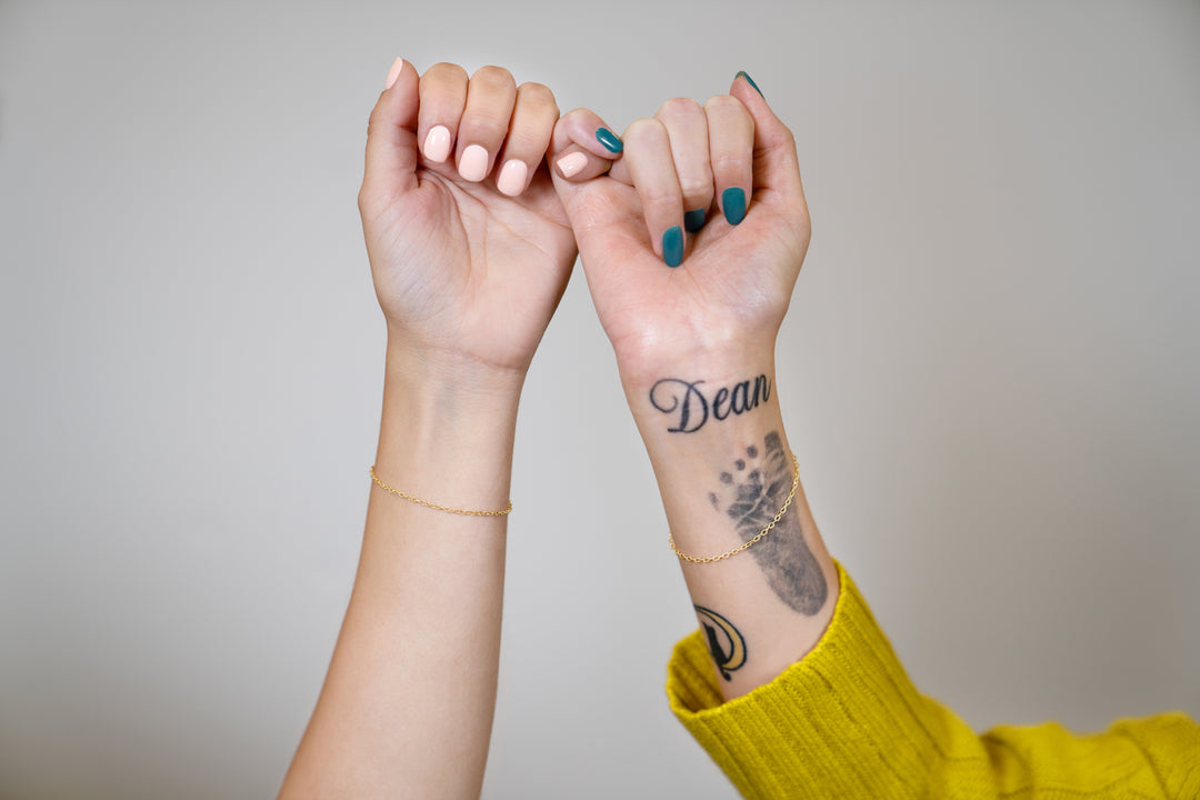 Two people's hands and forearms touching in a pinky promise pose against a plain light gray background. Both wear gold permanent bracelets.