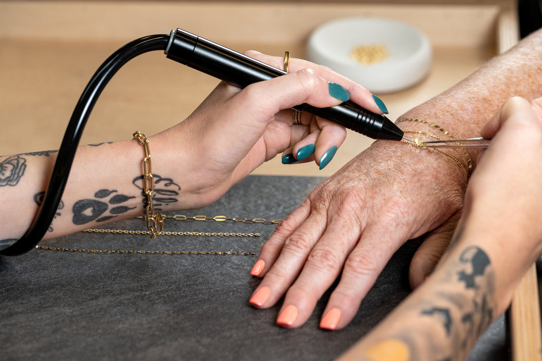A close-up of a jeweler's tattooed hands using a black handheld permanent jewelry device to attach three gold permanent bracelets. The jeweler has teal nail polish and black tattoos, and the scene is set on a dark gray table with a wooden background.