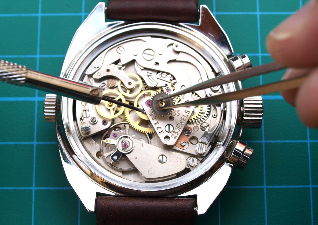 Close-up of a watch movement with a silver-toned case, brown leather strap, and visible internal gears and springs, being worked on with tweezers on a green grid cutting mat. A finger is visible holding the tweezers.