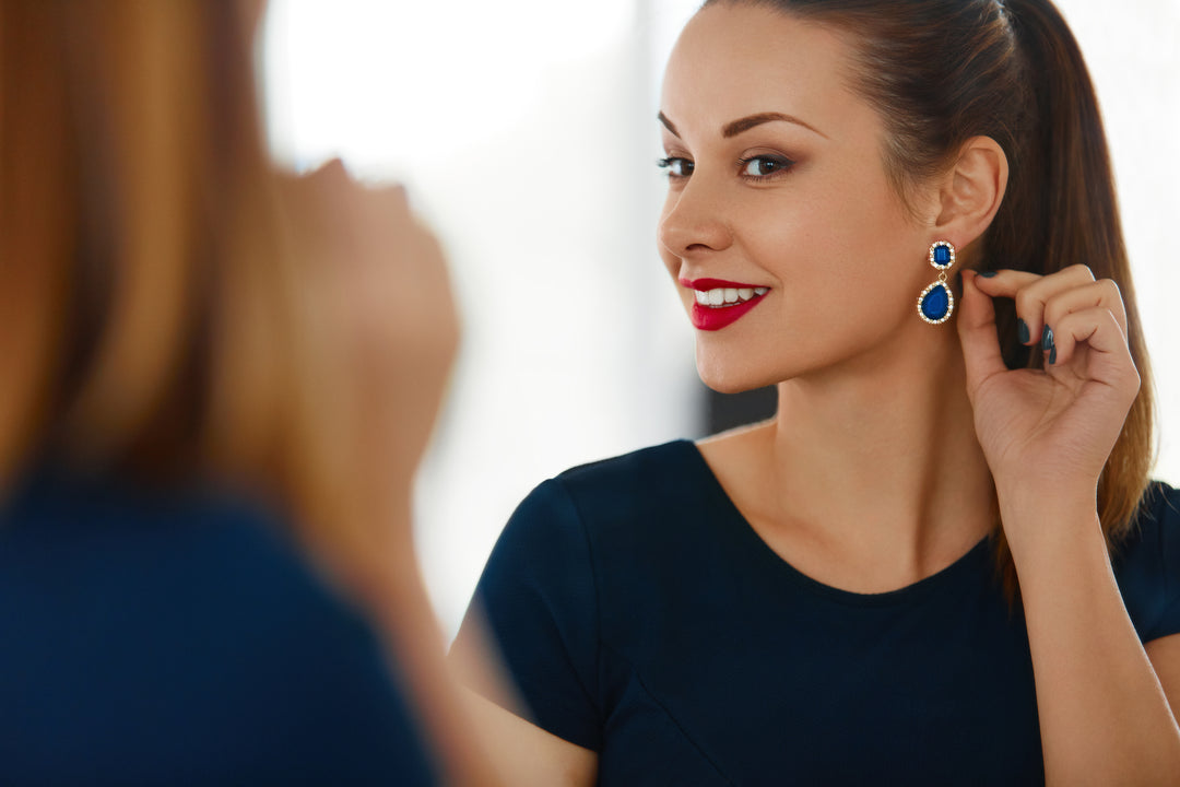 A lifestyle image of a woman wearing blue sapphire and diamond halo drop earrings and admiring them in a mirror. She wears red lipstick, blue nail polish, a blue short-sleeved shirt and has her long brown hair pulled back into a ponytail.