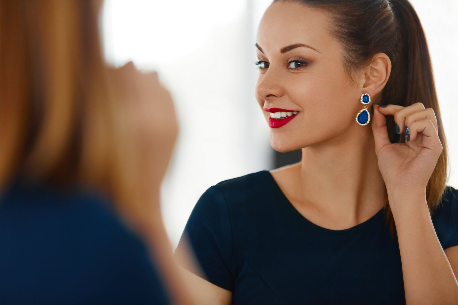 A lifestyle image of a woman wearing blue sapphire and diamond halo drop earrings and admiring them in a mirror. She wears red lipstick, blue nail polish, a blue short-sleeved shirt and has her long brown hair pulled back into a ponytail.