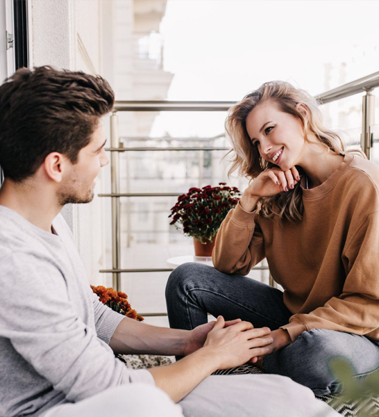 A couple talk about engagement rings and ring sizing on a balcony.