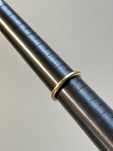 A plain gold band on a mandrel, a tapered metal bar with ring sizes, resting around US size 4.5.