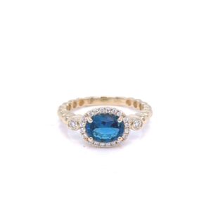 A 14 karat yellow gold ring with an oval faceted blue topaz in an east-west prong setting with round brilliant diamonds in the halo and one diamond in each shoulder of the band. The band has a polished bubble style design.