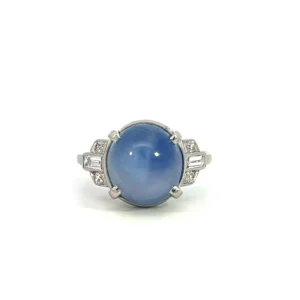 estate platinum ring set with 1 oval cabochon cornflower blur star sapphire weighing 9.62 carats accented on the shoulders with 4 single cut diamonds weighing 0.08 total carat weight and 2 baguette cut diamonds weighing 0.24 total carat weight. The ring is vintage from the 1920s.
