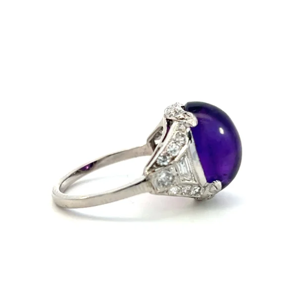 One estate vintage platinum amethyst ring from the 1920s featuring an oval cabochon dark amethyst measuring 12x10.40mm. The shoulders of the ring are accented with trapezoid diamonds, baguette diamonds, and round diamonds.