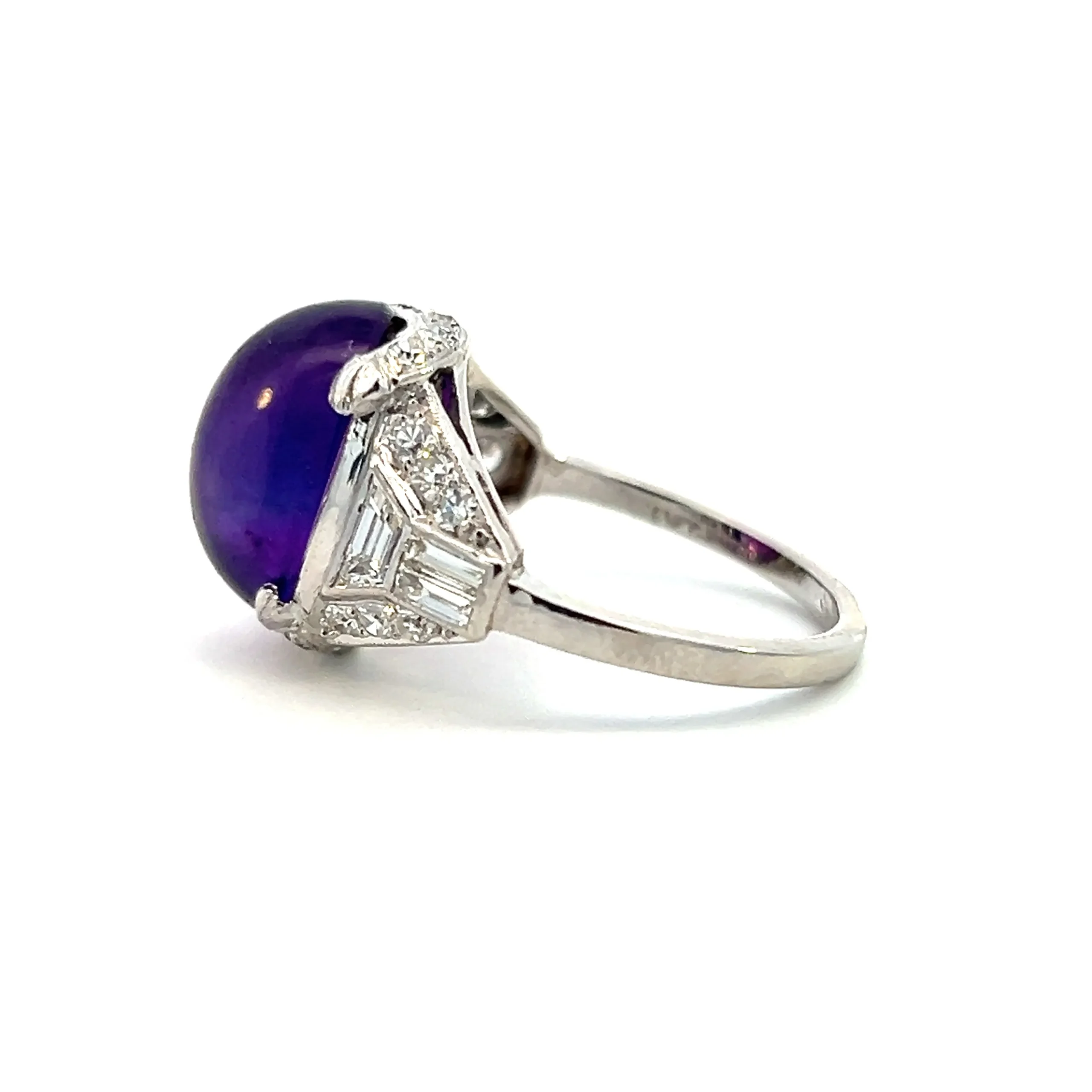 One estate vintage platinum amethyst ring from the 1920s featuring an oval cabochon dark amethyst measuring 12x10.40mm. The shoulders of the ring are accented with trapezoid diamonds, baguette diamonds, and round diamonds.