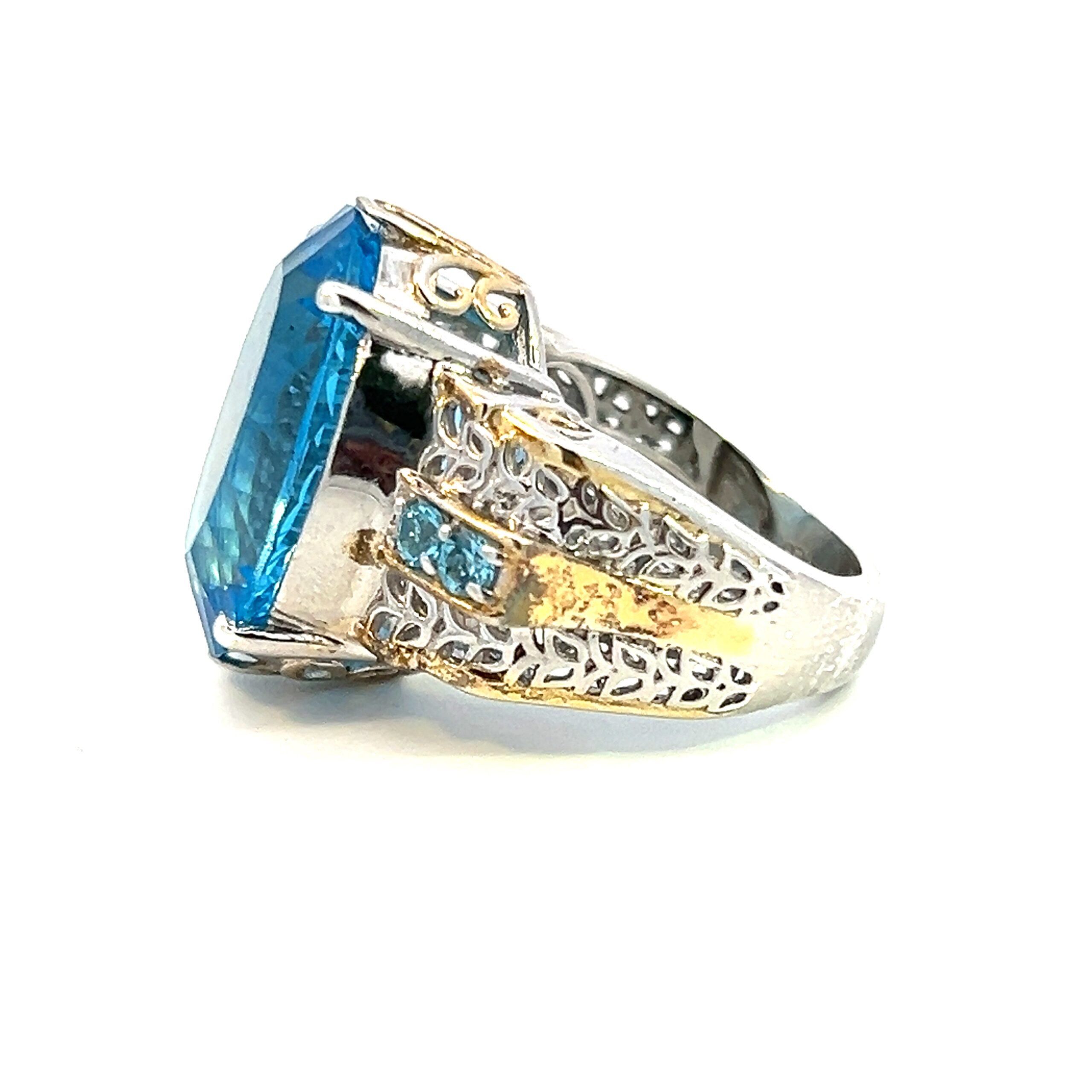 One estate two-tone blue topaz ring crafted from sterling silver with yellow gold-plated accents. The ring is set with a center 19x15mm oval blue topaz. The shoulders of the ring feature a center row of round blue topazes with nature-inspired filigree accents on both sides and nature-inspired filigree accents in the gallery area of the center setting. The gold plated accented are in the band only.
