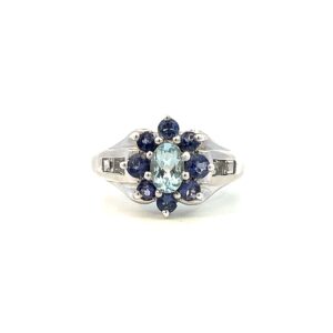 One estate tanzanite and blue topaz flower ring containing a center oval light blue topaz, 8 round tanzanite measuring 3mm each set as the petals around the blue topaz, and 2 square white sapphires in channel settings on both shoulders of the band.