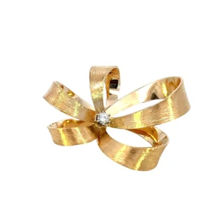 One estate 14 karat yellow gold diamond pin from the 1960s featuring a round brilliant diamond measuring 2.80mm and weighing 0.08 carat with H color and SI2 clarity in a six-prong setting in the center of an open-style bow with textured ribbons.