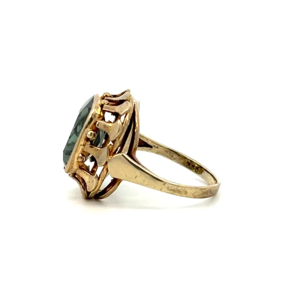 One estate 8 karat yellow gold gemstone ring containing a cushion-cut synthetic green spinel measuring 12x12.5mm in a bezel setting surrounded by an open-work gold frame. The entire top of the ring measures 18x17.5mm. Stamped "333" with a total weight of 4.23 grams. Vintage from the 1930s.