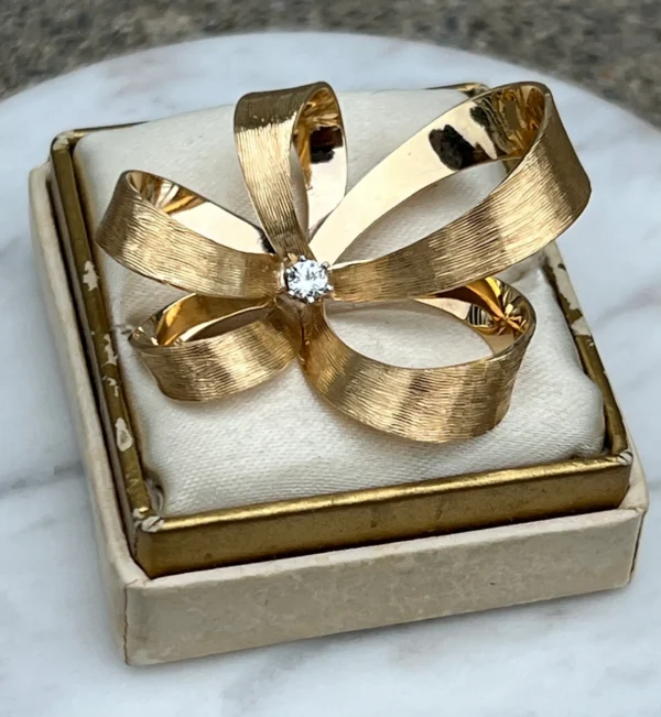 One estate 14 karat yellow gold diamond pin from the 1960s featuring a round brilliant diamond measuring 2.80mm and weighing 0.08 carat with H color and SI2 clarity in a six-prong setting in the center of an open-style bow with textured ribbons.